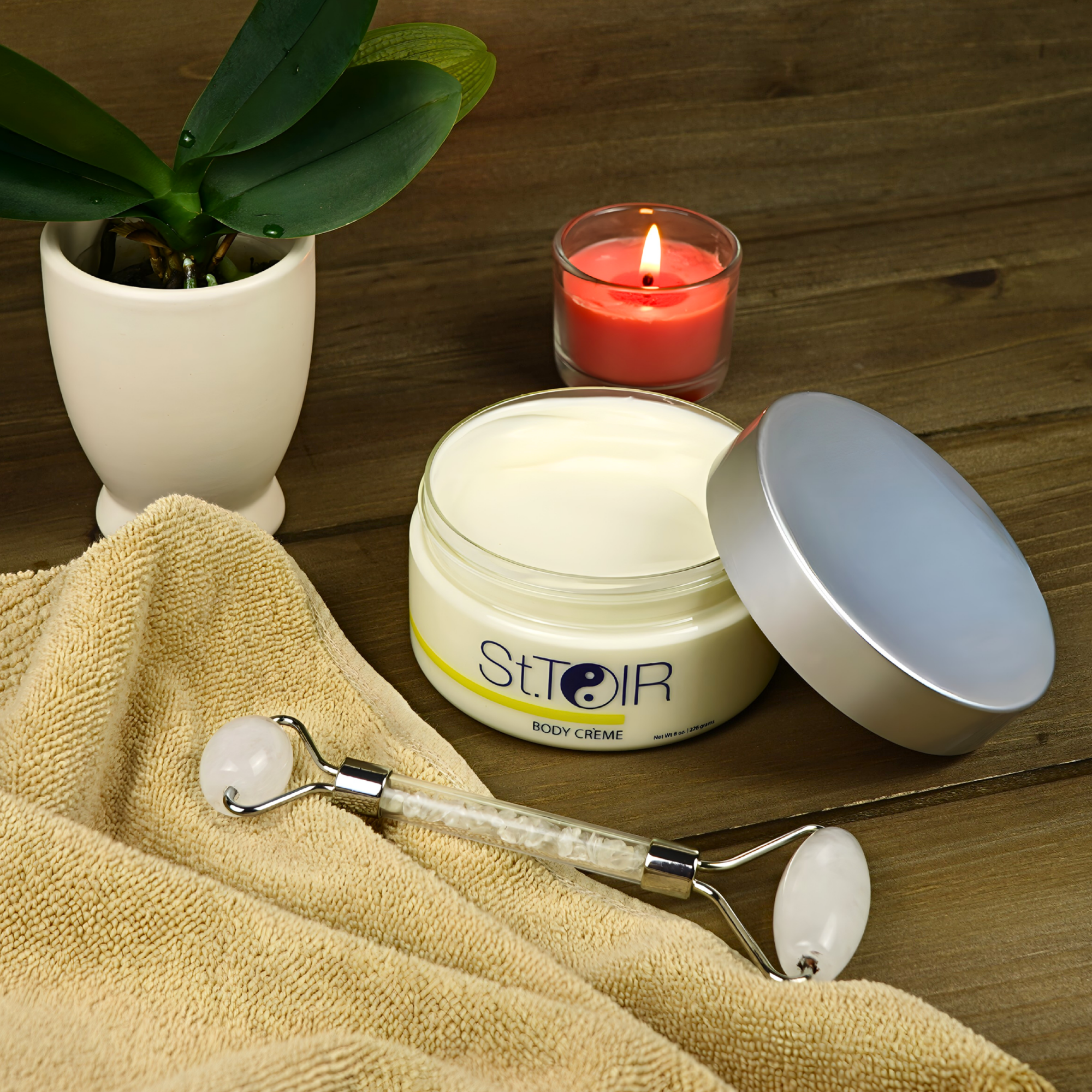 St. Ther's body crème jar with a roller on a towel next to a candle and plant.